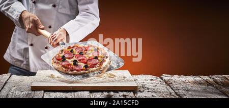 Cuisinier dans une pizzeria avec pizza salami maison affichée sur une palette métallique sur une surface farinée conseil en face de la lumière du feu dans le four dans une rusti Banque D'Images