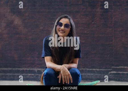 Belle jeune femme assise sur planche sur la rue portant des lunettes de soleil Banque D'Images