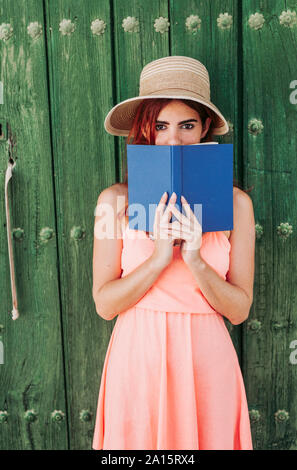 Portrait de jeune femme rousse se tenait devant la porte en bois vert couvrant la bouche avec un livre Banque D'Images