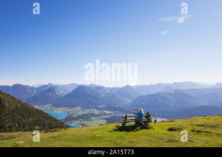 Vue arrière du senior male hiker photographing tandis qu'assis sur un banc en regardant les montagnes de Dachstein contre le ciel bleu Banque D'Images