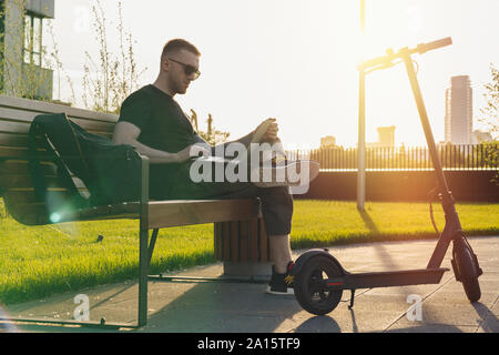 Jeune homme séduisant avec ordinateur portable et modern electric scooter de coup assis au parc contemporain au banc. et de travail indépendant ou business man workin Banque D'Images