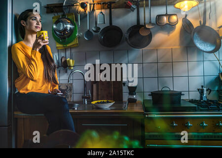Jeune femme assise sur le comptoir de la cuisine à la maison prendre un café Banque D'Images
