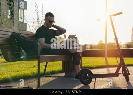 Jeune homme séduisant avec ordinateur portable et modern electric scooter de coup assis au parc contemporain au banc. et de travail indépendant ou business man workin Banque D'Images