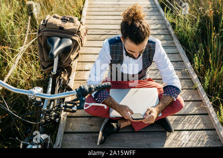 Bien habillée homme assis sur une passerelle en bois à la campagne à côté d'un vélo à l'aide d'un téléphone cellulaire et ordinateur portable Banque D'Images