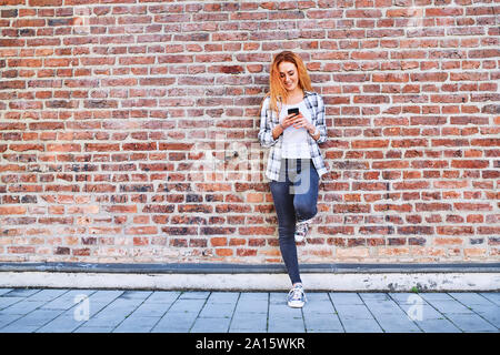 Jeune femme appuyée contre un mur de briques dans la ville lors de l'utilisation de smartphone Blackberry Banque D'Images