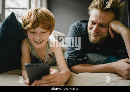 Portrait de l'heureux père et fils couché ensemble sur la table at cell phone Banque D'Images