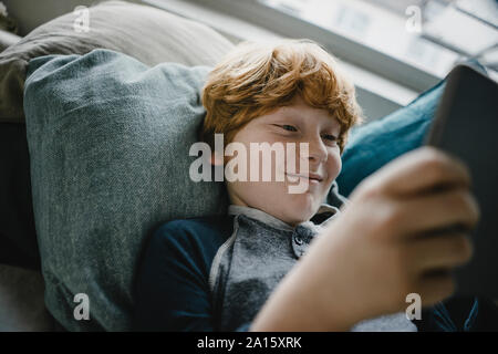 Portrait of smiling redheaded boy lying on sofa using digital tablet Banque D'Images