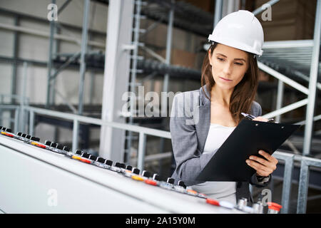 Woman wearing hard hat sur le panneau dans une usine à prendre des notes Banque D'Images