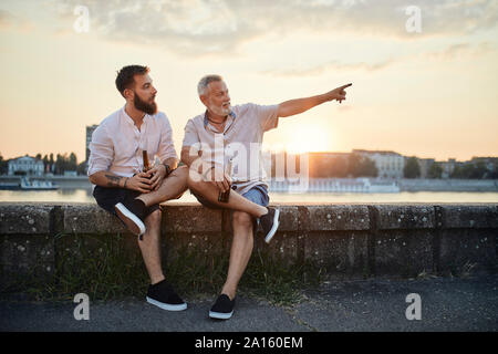 Père et fils adultes assis sur un mur au bord du fleuve au coucher du soleil de boire une bière Banque D'Images