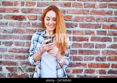 Cheerful young woman using smartphone pendant que appuyé contre un mur de briques Banque D'Images