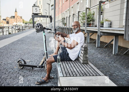 Portrait d'homme mûr avec l'E-scooter se détendre sur un banc en été en utilisant un téléphone cellulaire, Cologne, Allemagne Banque D'Images