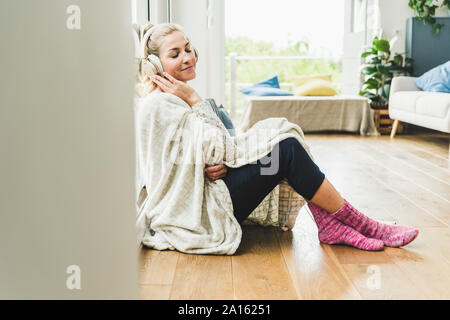 Femme enveloppée dans une couverture assis à la fenêtre à la maison écouter de la musique avec des écouteurs Banque D'Images