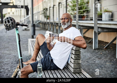 Portrait d'homme mûr avec l'E-scooter et le téléphone portable assis sur un banc en été Banque D'Images