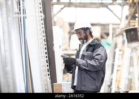 Jeune homme avec cell phone wearing hard hat travaillant dans un entrepôt Banque D'Images