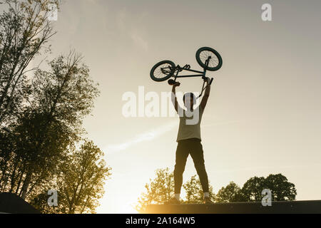 Jeune homme soulevant vélo BMX au skatepark au coucher du soleil Banque D'Images