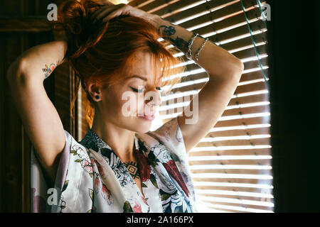 Red-haired femme tatouée avec les yeux fermés à la maison près de la fenêtre Banque D'Images