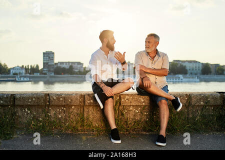 Père et fils adultes assis sur un mur au bord de la rivière parle Banque D'Images