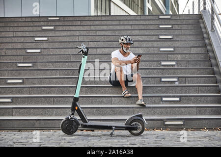 Homme mûr avec l'E-Scooter sitting on stairs using cell phone Banque D'Images