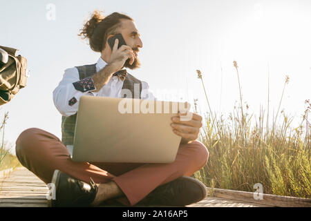 Bien habillée homme assis sur une passerelle en bois à la campagne avec ordinateur portable et téléphone cellulaire Banque D'Images