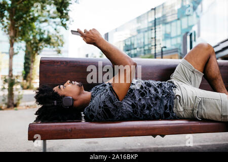 Homme couché sur un banc à l'écoute de la musique avec des écouteurs lors de l'utilisation de téléphone mobile, Barcelone, Espagne Banque D'Images