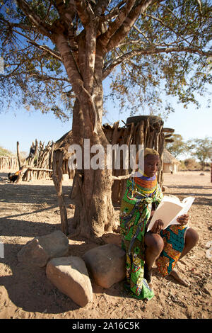 Femme muhila traditionnel, assis sous un arbre, lecture, livre Kehamba, Chibia, Angola Banque D'Images