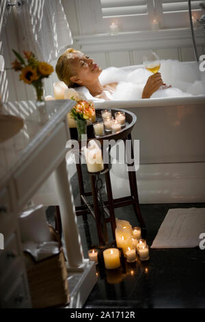 Mid-adult woman relaxing dans un bain avec un verre de vin et entouré de bougies. Banque D'Images