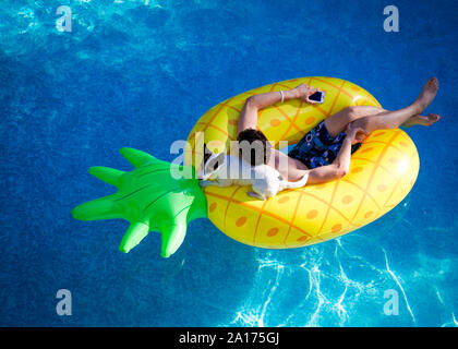 Teenage boy holding sur un téléphone mobile et son petit chien détente sur une journée d'été, à flot sur un ananas gonflable dans une piscine extérieure. Banque D'Images