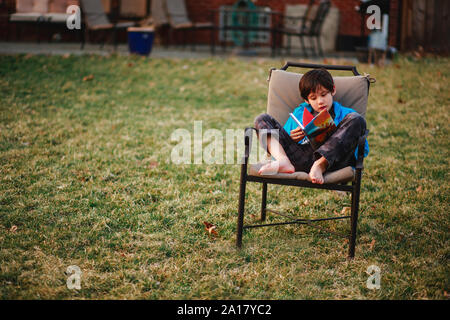 Un petit enfant est assis sur une chaise, pieds nus dans le jardin en lisant un livre au printemps Banque D'Images