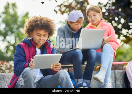 African boy avec des cheveux bouclés assis à l'extérieur et à l'aide de tablet pc avec d'autres enfants à l'aide d'un coffre à l'arrière-plan Banque D'Images