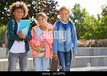 Portrait of happy group of school children smiling at camera while Standing together en plein air avec des livres et des planches à roulettes Banque D'Images