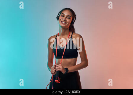 Sport avec musique. Jeune femme sportive dans un casque tenant une corde de saut et regardant la caméra avec le sourire tout en se tenant debout contre un arrière-plan coloré. Concept sport et fitness. Mode de vie actif Banque D'Images
