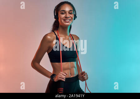 La musique est ma motivation. Jeune femme sportive dans un casque tenant une corde de saut sur les épaules et regardant l'appareil photo avec le sourire tout en se tenant contre un arrière-plan coloré. Concept sport et fitness. Mode de vie actif Banque D'Images