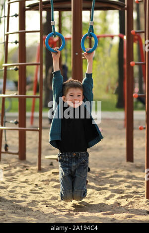 Happy smiling boy hanging on gymnastic rings sur l'aire de jeux extérieure. Portrait candide full size Banque D'Images