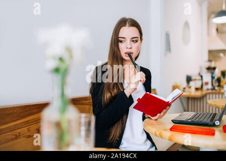 Les jeunes concentrated businesswoman holding son calepin et stylo, assis en face de l'ordinateur portable dans un café. Banque D'Images