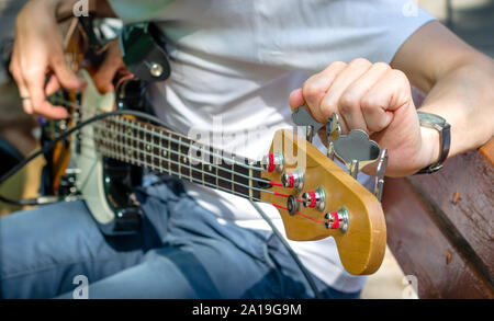 Musicien de rue dans un T-shirt blanc et jean bleu tunes une 4-cordes guitare basse assis sur un banc dans un parc sur une journée ensoleillée. Banque D'Images