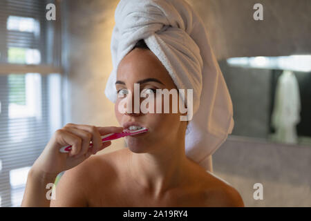 Jeune femme brune se brosser les dents dans une salle de bains avec ses cheveux dans une serviette Banque D'Images