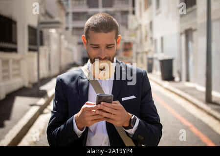 Jeune homme professionnel de manger un beigne et à l'aide du smartphone sur la rue Banque D'Images