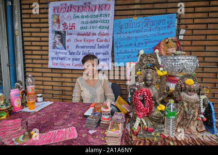 Une femme Fortune Teller avec ustensiles de son métier et des statues de divinités hindoues, assis en face d'un temple hindou à Silom Rd., Bangkok, Thaïlande Banque D'Images