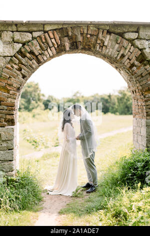 Beau couple, femme chinoise en robe de mariage de luxe et l'homme en costume gris, standing in old vintage stone arch, à l'extérieur près du château Banque D'Images