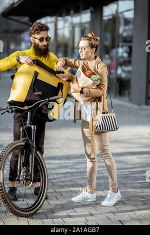 Courrier mâle sur un vélo offrant des aliments frais à l'aide de sac thermique à une jeune femme d'affaires à l'extérieur, femme un engagement sur un smart phone Banque D'Images