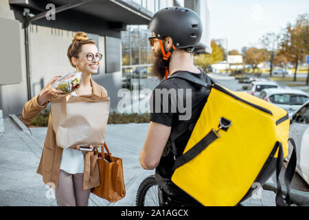 Courrier mâle la livraison de nourriture sur un vélo, avec un sac à lunch à emporter frais d'une jeune femme d'affaires à l'extérieur. Livraison de nourriture restaurant concept Banque D'Images