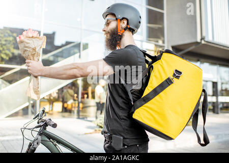 Jeune et joyeuse attente de courrier avec des fleurs pour un client près du bâtiment à l'extérieur, la prestation d'une bicyclette frais accessoires à l'aide de sac thermique Banque D'Images