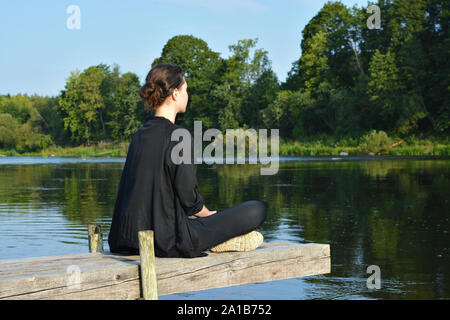 Jeune fille dans un survêtement et une veste noire est assis en tailleur sur la passerelle sur la rive et est titulaire d'une session de méditation. Banque D'Images