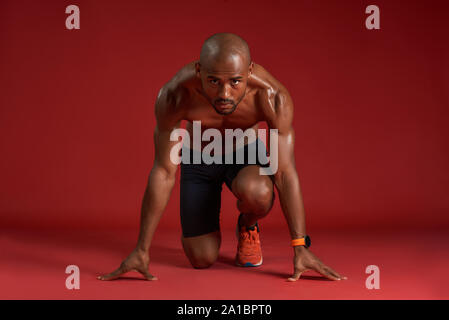 Portrait de jeune homme africain dans les vêtements de sport s'exécuter tout en restant assis sur fond rouge isolé. De fitness et de work out concept. Plan horizontal Banque D'Images