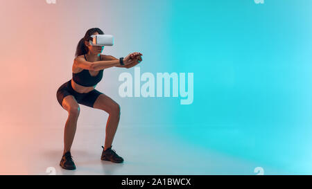 Toute la longueur de jeune femme sportive dans les vêtements de sport faisant s'accroupir tout en portant des lunettes de réalité virtuelle. Studio shot. La technologie de l'avenir Banque D'Images