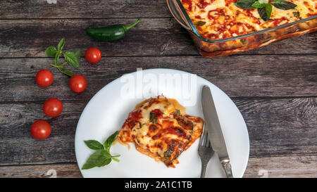 L'aubergine parmigiano, un morceau sur une assiette avec une fourchette et couteau, à côté du bac avec les aubergines, tomates cerises Banque D'Images