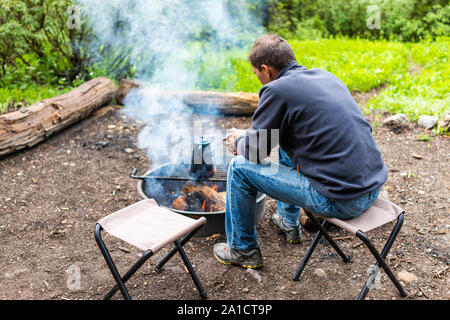 Camping avec man sitting on chair réchauffement par la fumée du foyer et théière sur grill à Albion, bassin d'été dans les montagnes Wasatch de l'Utah en soir Banque D'Images