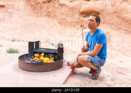 Jeune homme soufflant carbonisés rôti de refroidissement des guimauves sur feu de camp au camping grill dans paysage de désert en parc d'état de Goblin Valley Banque D'Images