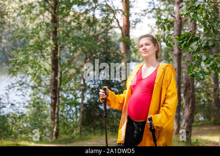 Portrait of young pregnant woman wearing raincoat en été permanent forest holding bâtons appréciant la nature - Grossesse en santé concept de vie Banque D'Images
