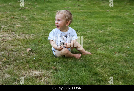 Une malheureuse petite boueux bébé garçon jouant dans le jardin portant un t-shirt blanc sale haut sitting on grass s'assied et pleure Banque D'Images